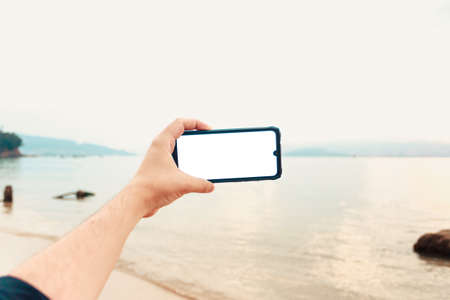 A young male hand grabbing a phone with copy space in horizontal with the ocean as the backgroundの写真素材