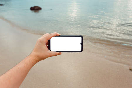 A young male hand grabbing a phone with copy space in horizontal with the sand and the sea of the beach as the backgroundの写真素材