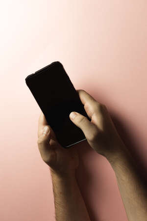 A top view of two young hands using a mobile phone with copy space and a black screen over a pink pastel backgroundの写真素材