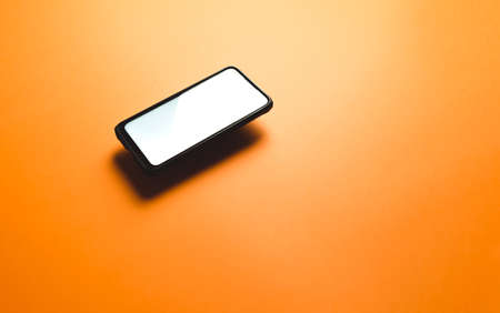 A minimalistic mock up flat image design with a floating mobile phone with copy space and white scree to write over it over a flat orange backgroundの写真素材