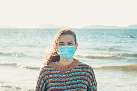 A young woman on a sweater on the beach using a surgical mask during a bright day with an island as the backgroundの写真素材