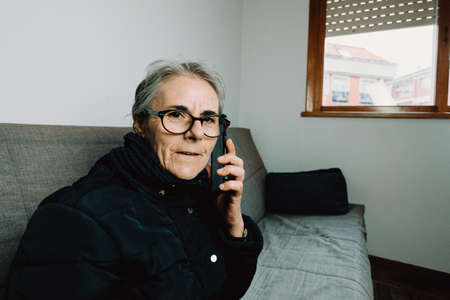 An old woman having a phone call while smiling to the camera in a living roomの写真素材