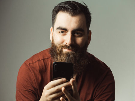 A young hipster style man with a beard and modern haircut smiling while looking at camera with his phone screen with copy spaceの写真素材