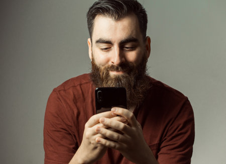 A young hipster style man with a beard and modern haircut smiling while watching his phone screen with copy spaceの写真素材