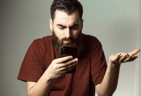 A young hipster style man with a beard and modern haircut angry while looking at his phone screen with copy spaceの写真素材