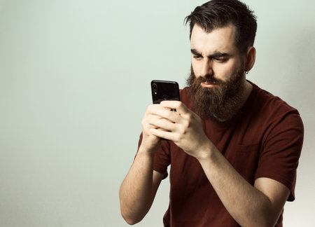 A young hipster style man with a beard and modern haircut angry while looking at his phone screen with copy spaceの写真素材