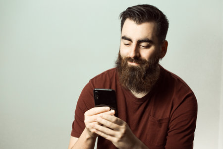 A young hipster style man with a beard and modern haircut smiling while watching his phone screen with copy spaceの写真素材