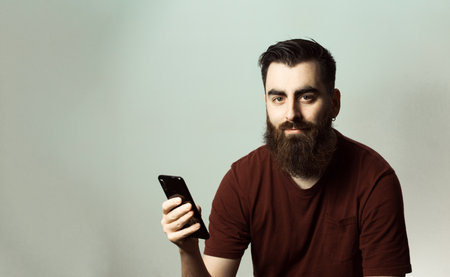 A young hipster style man with a beard and modern haircut smiling while looking at camera with his phone screen with copy spaceの写真素材