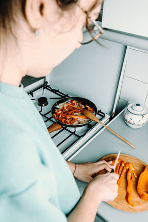 A woman cutting some pepper over a wooden round plate with a knife while cooking with the pan in the kitchenの写真素材