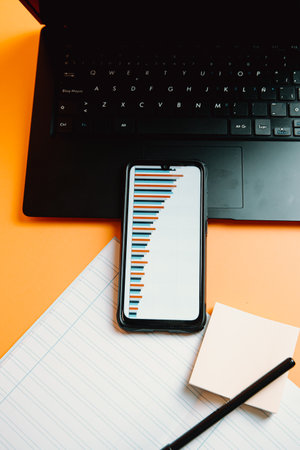A phone with statistics over a orange background with laptop and notebook with financial conceptの写真素材
