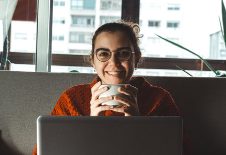 A young woman smiling in front of his laptop in the couch while holding a cup of coffeeの写真素材