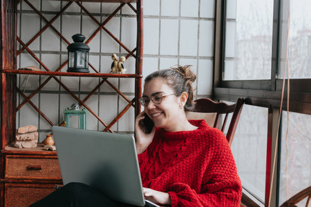A young woman smiling while working on his laptop and making a call on an indie modern houseの写真素材