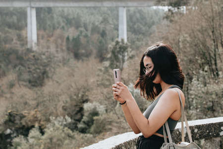 Young woman on sport clothes and sunglasses looking from a balcony with copy space during a sunny day, checking his phone and making photosの写真素材