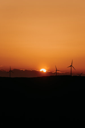 Silhouettes of some wind mills on the top of a mountain during a super orange sunset with copy space peacefulの写真素材