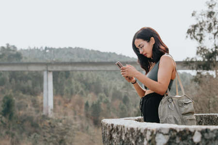 Young woman on sport clothes and sunglasses looking from a balcony with copy space during a sunny day, checking his phone and making photosの写真素材