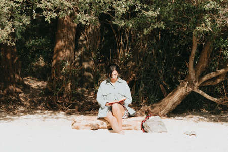 Young Moroccan woman on modern clothes sitting on the beach reading a book during a sunny day with copy space inspirational and relax theme with colorful tonesの写真素材