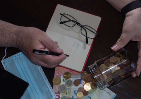 counting the coins and notes saved in a glass jar and noting them in the notebook with the calculator, after the mask confinement and economic crisis by the covid1の写真素材
