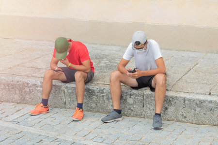 two young teenagers sitting on the stone pavement playing and chatting on social networks with their...