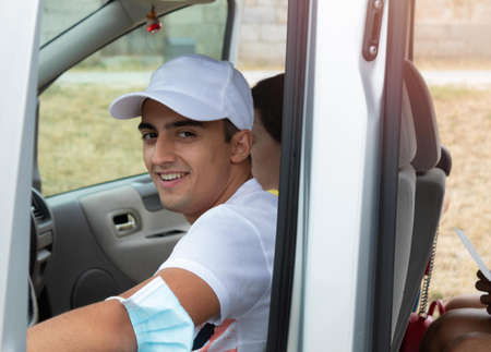 young teenage boy smiling in the car, ready to drive a trip with the family and wearing the health mask on his elbow on a summer dayの写真素材