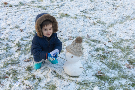 Little boy in a coat, gloves and blue boots crouching down with his snowman on a cold winter dayの写真素材