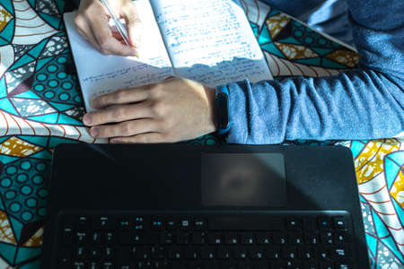 selective close-up focus of a student's hand with a sports watch taking notes in a notebook and studying with a black laptop computerの写真素材