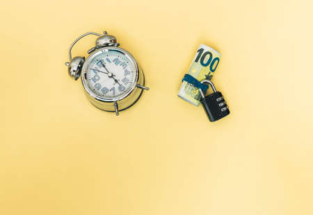 selective approach of several rolled banknotes held with an elastic band and a closed padlock and an analog clock showing the time with copy space and yellow background concept time is moneyの写真素材