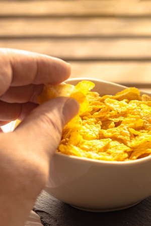 Selective focus of a person picking up cornflakes for breakfast with his hand from a full bowl on a wooden table.の写真素材