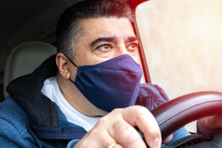 Middle-aged man with protective mask against airborne virus driving very attentive and tense in...