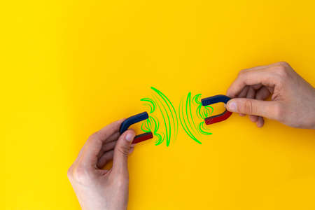 detail of hands holding two blue and red horseshoe-shaped magnets showing how equal poles repel each other with lines indicating this on yellow background and copy space concept breakage disagreementの写真素材