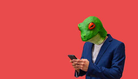 man with googly-eyed frog mask texting or watching news with smart cell phone on red background with copy spaceの写真素材
