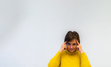 middle-aged woman in yellow sweater trying with head and hands to read mind and worried on white...