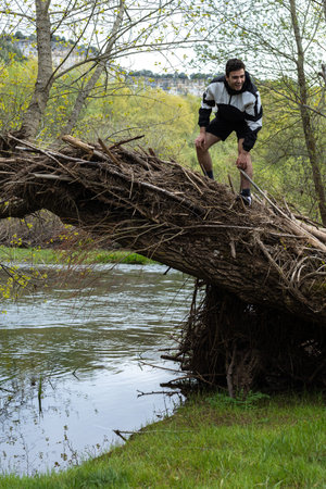 young man in sportswear and shorts climbing on a fallen tree trunk laughing after having achieved...