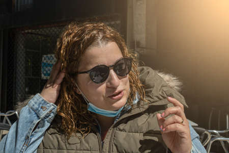 woman with curly hair and sunglasses with her mask pulled down for having a snack on the terrace of a restaurant bar on a sunny day with copy spaceの写真素材