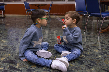 a child sitting on the floor of a cafeteria playing with a pompero that blows soap bubbles through it, amusing another child with pitt hopkins syndrome.の写真素材