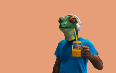person with frog mask with a glass jar with fruit juice and a straw to drink and refresh while listening to music through headphones on orange background with copy spaceの写真素材