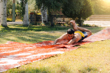 teenage boy in a bathing suit jumping down a soap and water slide or pool with his arms outstretched to slide down the garden and cool off on a hot summer day.の写真素材