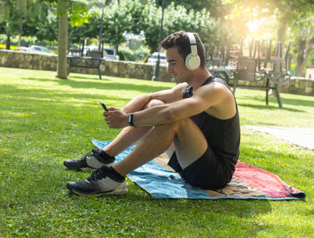 young Caucasian male athlete sitting on a towel in sportswear listening to cell phone music through   headphones on a sunny summer day.の写真素材