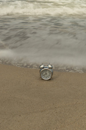 vertical photo of an analog clock on the shore of the beach with the silky waves at dawn on a summer...