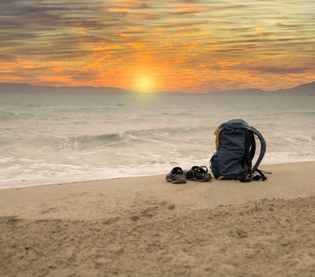adventurer's backpack with flip flops on the sandy shore on the beach with sunrise over the sea with copy spaceの写真素材