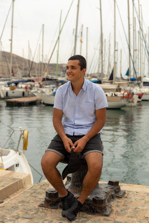Caucasian teenager in smiling shorts sitting on top of a mooring bollard in the harbor with ships in...