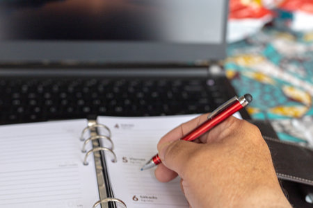 detail of a young entrepreneur writing down events in a paper diary in front of a computer in the organizations officeの写真素材