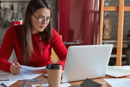 Horizontal shot of busy female worker with dark hair, wears spectacles, collects information for financial report, checks information from electronic version, focused into laptop, sits at kitchenのeditorial素材