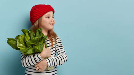 Cute small child in red headgear and striped jumper, carries green salad, looks away, wants to make fresh salad, keeps to healthy eating, stands sideways against blue background with free spaceの写真素材