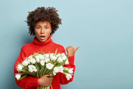 Shocked puzzled woman with Afro hairstyle, points away with thumb, carries beautiful white spring flowers, shows way to florist, amazed by high price on tulips, wears red sweater, poses indoorの写真素材
