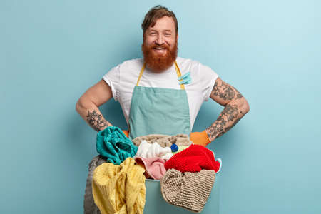 Positive red haired housekeeper glad to finish domestic work, keeps hand on waist, wears casual t shirt and apron, prepares for washing linen, poses over blue background. Laundry day conceptの写真素材
