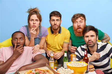 Fascinated friends stare at screen, watch horror film together at home, spend weekend evening in domestic atmosphere, drink beer and eat fast food, have scared faces. People, spare time, friendshipの写真素材