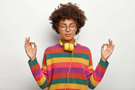 Foto de Relaxed beautiful lady with Afro hairstyle meditates indoor, keeps eyes closed, feels satisfied lo listen pleasant music in headphones, wears spectacles, earrings, striped jumper. Body language - Imagen libre de derechos
