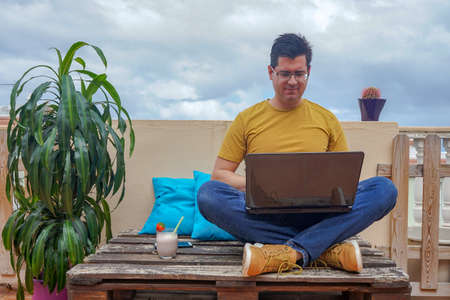 Man working from the terrace of his house. The man is drinking a strawberry smoothie. He is working with his computer telematically.の写真素材