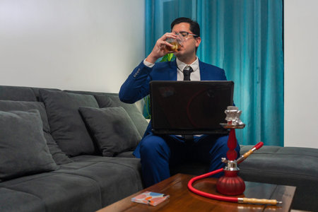 Young man sitting on a sofa. He is wearing a blue suit. He is working on his laptop and drinking a...