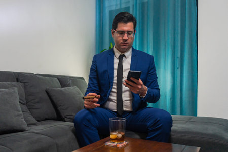 Young man in a blue suit sitting on a sofa. He is smoking a cigar and drinking a glass of Whiskey....